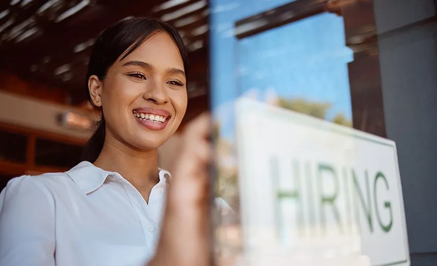 A restaurant worker smiling and holding a sign that says "hiring"
