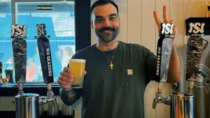 Theo Castillo stands behind the bar, gripping a beer and flashing a victory sign with two fingers on his other hand.
