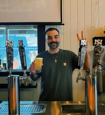 Theo Castillo stands behind the bar, gripping a beer and flashing a victory sign with two fingers on his other hand.