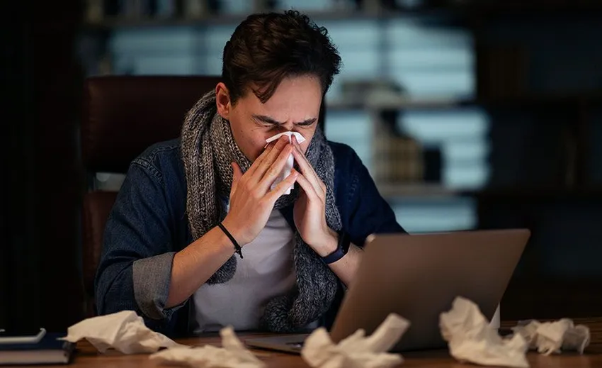 Unhealthy sick young man with scarf around his neck employee working late at office, guy sitting at workdesk, looking at laptop screen and sneezing, using napkin