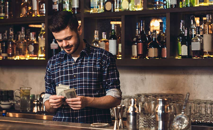A smiling bartender stands behind the bar and counts the money