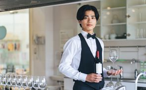 A young Asian male sommelier holding a bottle of wine with his right hand and a wine glass by its stem with his left hand