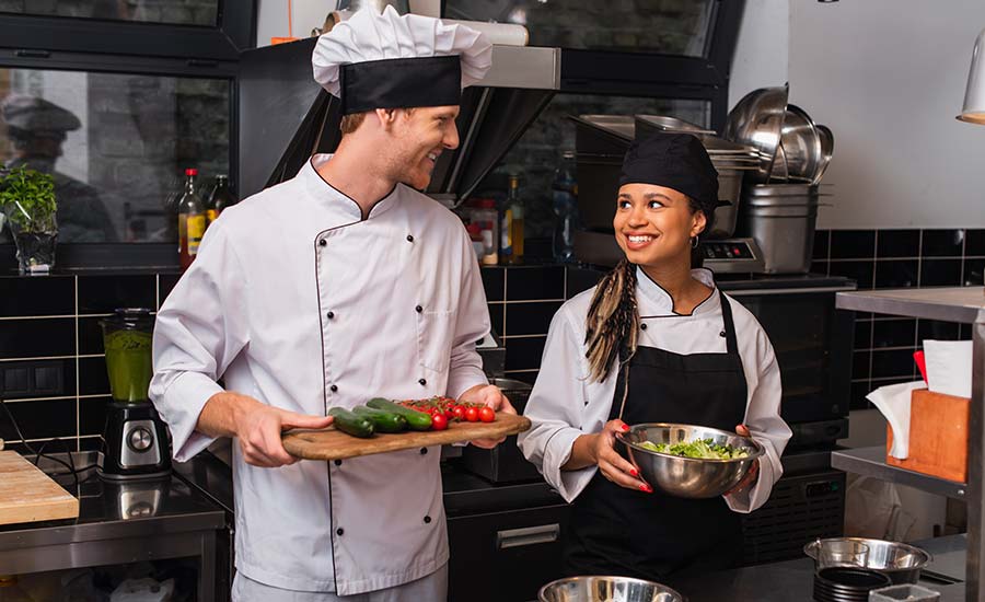 Sous chef salary: a male ad female cook are smiling and looking at each other, holding dishes in full uniform, in a restaurant kitchen.
