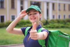 portrait of young beautiful happy courier teenager girl, food delivery woman with green thermo box for food delivering food outdoors in the yard at summer day in cap and uniform