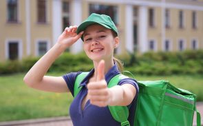 portrait of young beautiful happy courier teenager girl, food delivery woman with green thermo box for food delivering food outdoors in the yard at summer day in cap and uniform