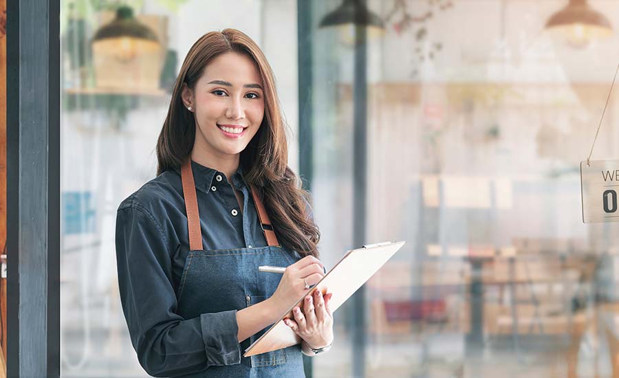 Beautiful Asian young hostess in apron holding clipboard and standing in front of the door of cafe with open sign board
