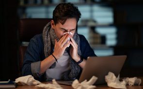 Unhealthy sick young man with scarf around his neck employee working late at office, guy sitting at workdesk, looking at laptop screen and sneezing, using napkin