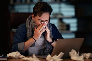 Unhealthy sick young man with scarf around his neck employee working late at office, guy sitting at workdesk, looking at laptop screen and sneezing, using napkin