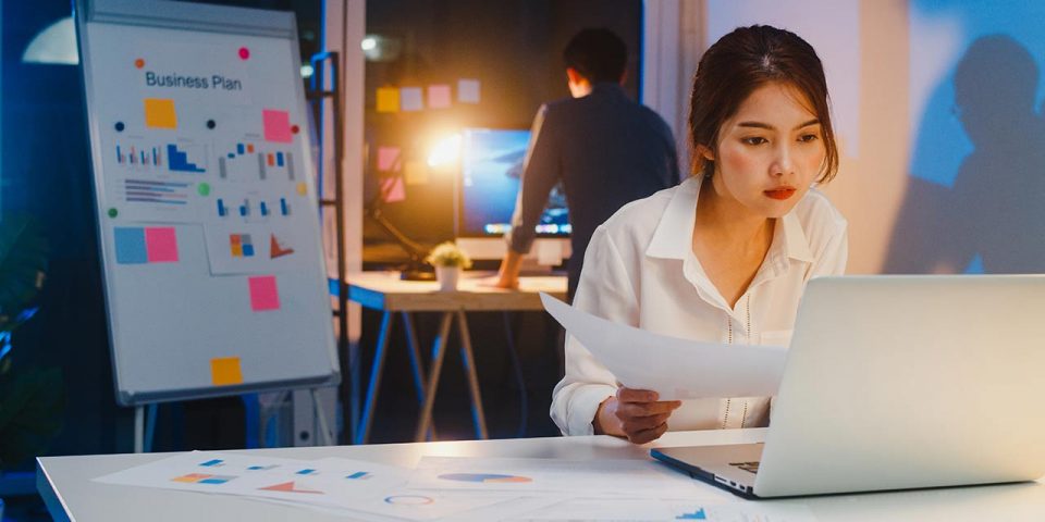 A girl in front of her laptop working overtime with a guy in the background