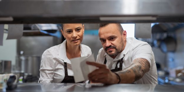 A male food expeditor and a female kitchen manager discussing a customer's order on a sheet of paper