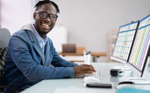 A male Black management analyst smiling while sitting in front of a desktop showing a spreadsheet