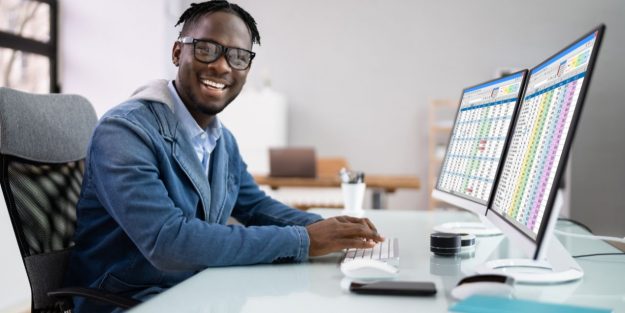 A male Black management analyst smiling while sitting in front of a desktop showing a spreadsheet