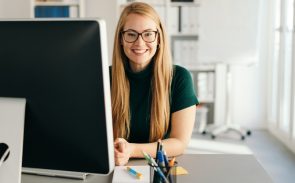 A female HR clerk with long hair and glasses smiling while sitting in front of her monitor