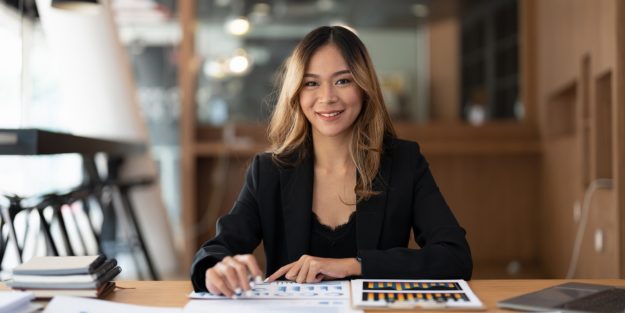 A female Asian payroll analyst smiling with her right hand on the desk