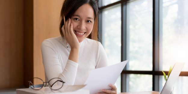A female accounts receivable clerk smiling with her left hand on her face and holding a sheet of paper on her right hand
