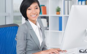 A short-haired female Asian administrator smiling while sitting on her desk in front of her monitor