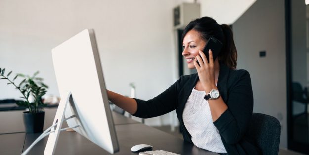 A sales administrator holds a phone to her ear while facing a monitor