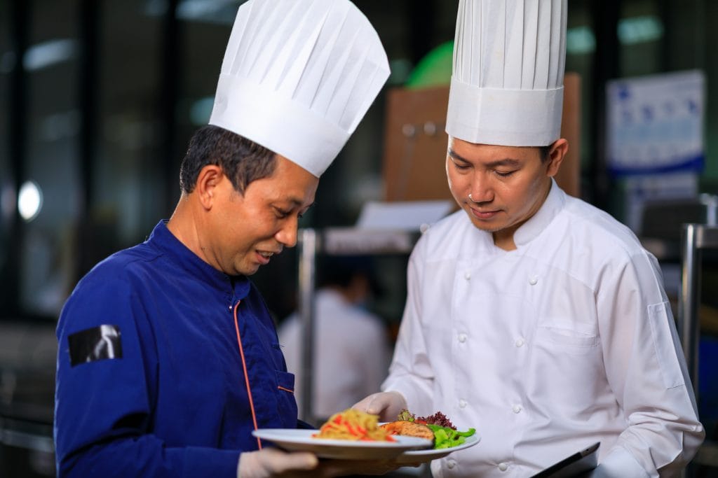 A male Executive Chef wearing blue discussing a dish with his Sous Chef