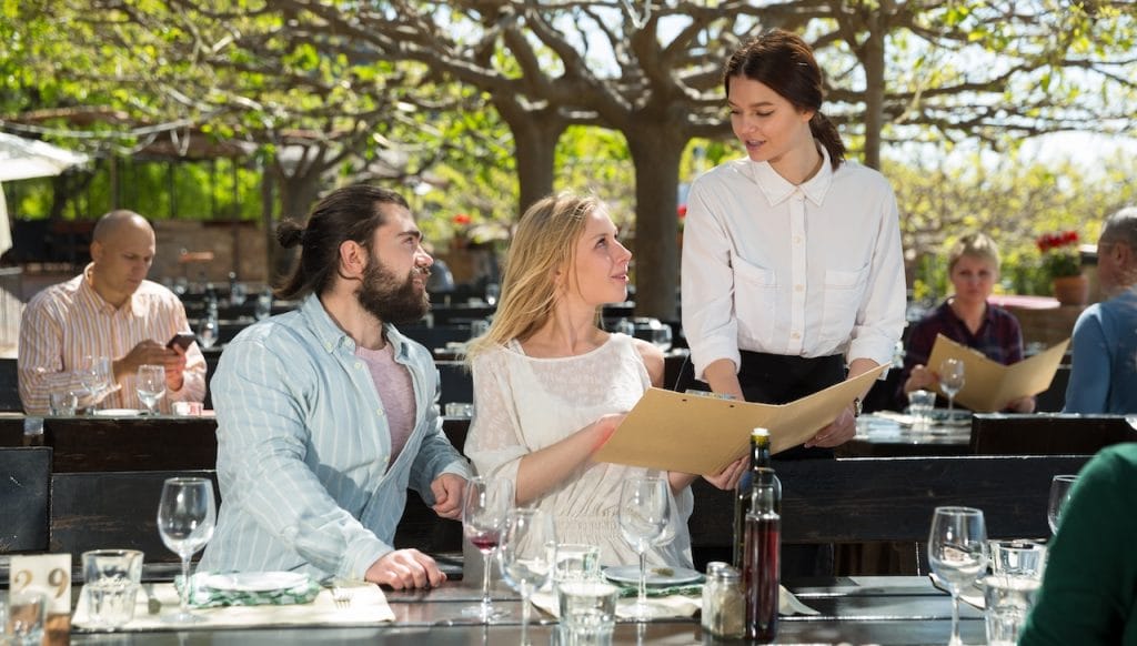 Charming young waiter and couple at open-air restaurant summer