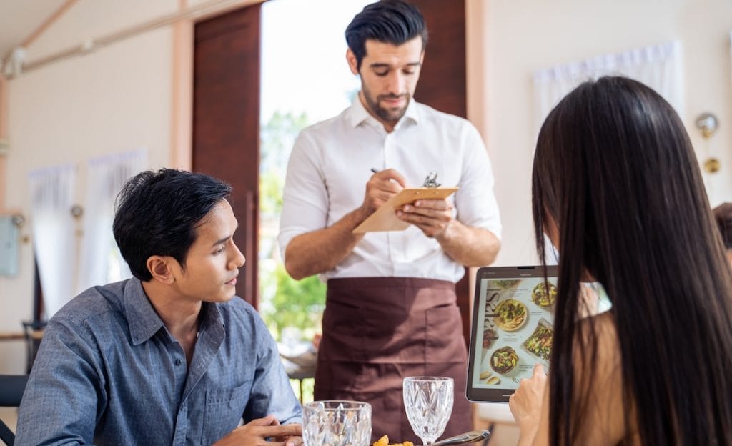 Caucasian waiter receiving order serving from customer in restaurant. Attractive server service man working, taking note, writing order from consumer with pen at table in dining room with happiness